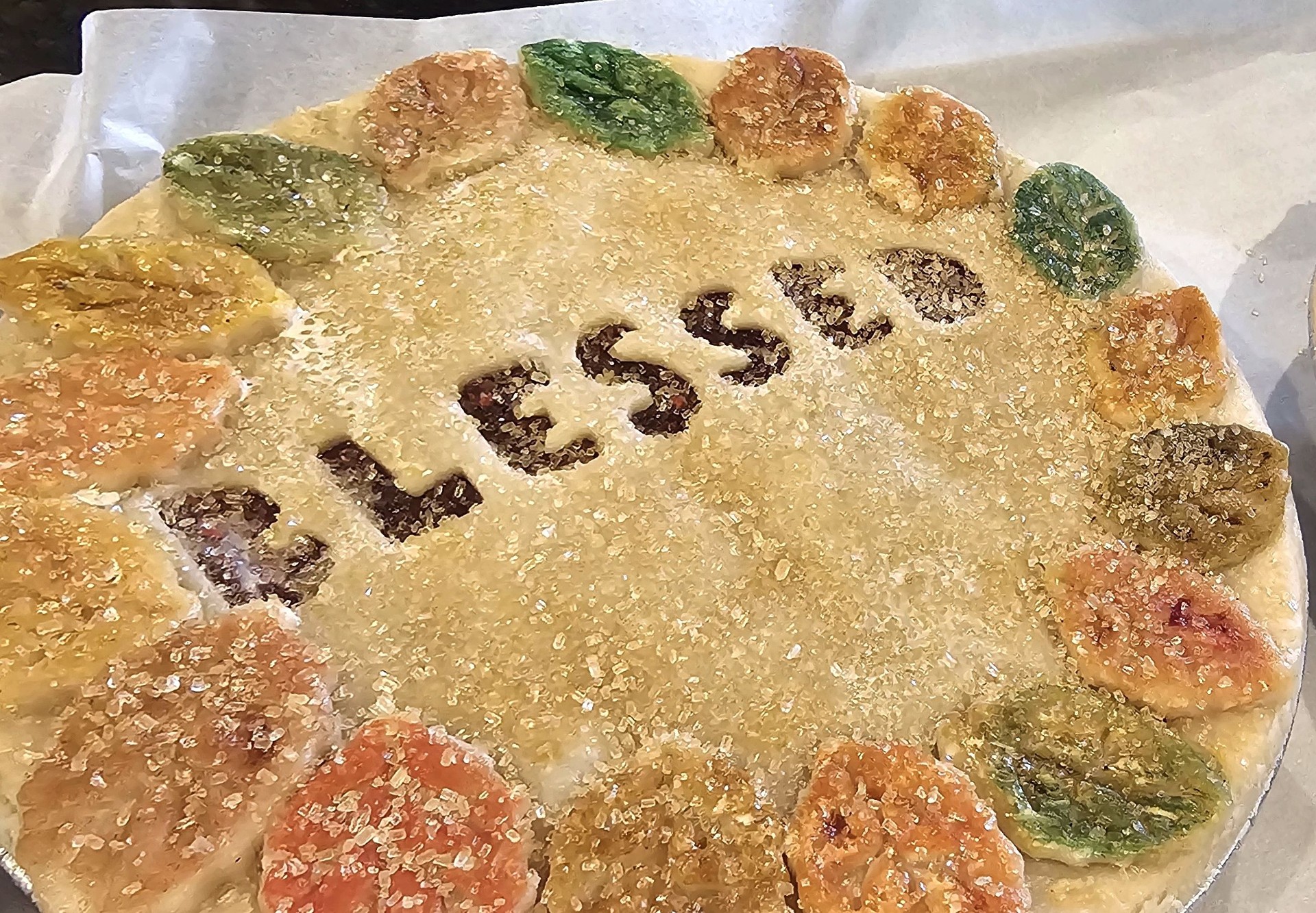 Round pie decorated with a sugared crust, colorful leaf-shaped accents, and 'BLESSED' stenciled in the center