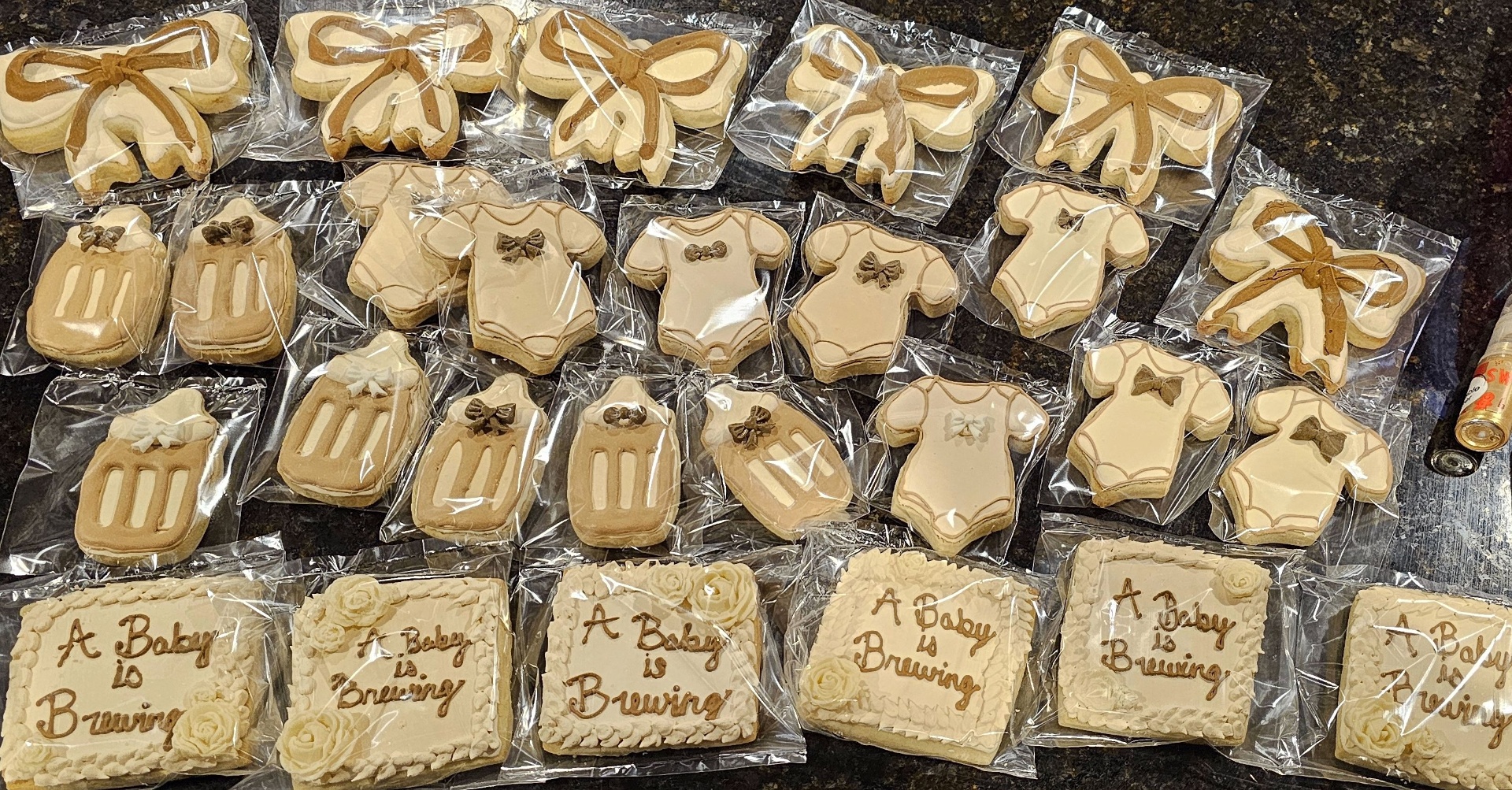 Neutral-toned baby shower cookies — bows, onesies, booties, and 'A Baby is Brewing' plaques